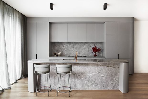 Contemporary minimalist kitchen featuring a large sculptural stone island with integrated seating, pale grey handleless cabinetry, a matching stone splashback, timber flooring, sheer full-height curtains, and brass tapware. The softly neutral palette and streamlined detailing create a refined, calm interior.
