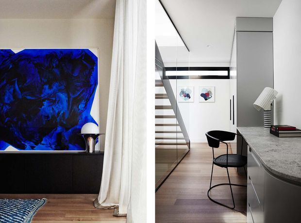 Contemporary open-plan interior with a sculptural staircase, grey built-in cabinetry and stone countertop, black dining chair, timber flooring, and a bold blue abstract artwork beside sheer curtains. The space is bright, refined, and minimalist with strong black-and-white contrast and warm timber accents.
