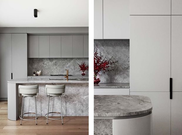 A refined contemporary kitchen featuring pale grey full-height cabinetry, a large stone island with a rounded end, and two white upholstered bar stools. The kitchen includes a textured grey stone splashback and matching island surfaces, slim black hardware and fixtures, warm timber flooring, and restrained red floral styling.