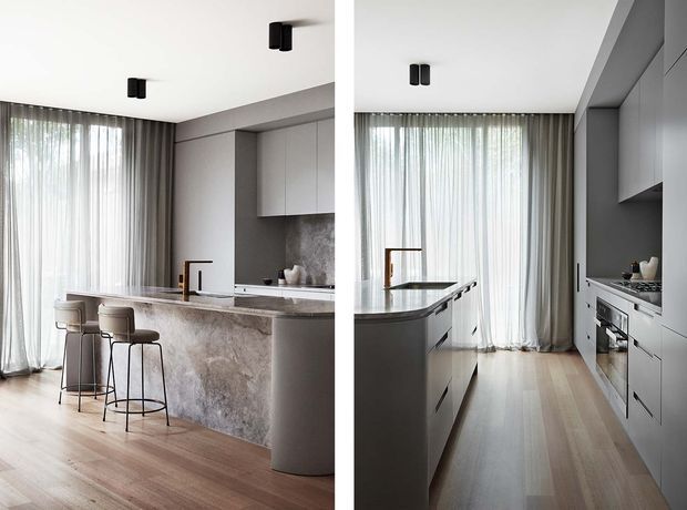 Split-view image of a refined contemporary kitchen, showing a large stone island with rounded end, integrated cabinetry, built-in appliances, a dark stone or marble splashback, timber flooring, and full-height sheer curtains over glazed doors or windows. The palette is light and neutral with warm wood flooring and subtle dark accents.