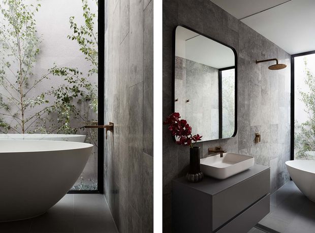 A refined contemporary bathroom with a freestanding white bathtub positioned beside full-height glazing and leafy planting outside. The room features grey concrete-look wall and floor finishes, a dark floating vanity with a white vessel basin, a rounded rectangular black-framed mirror, and warm bronze/copper-toned tapware and shower fittings. Natural light creates a calm, modern atmosphere.