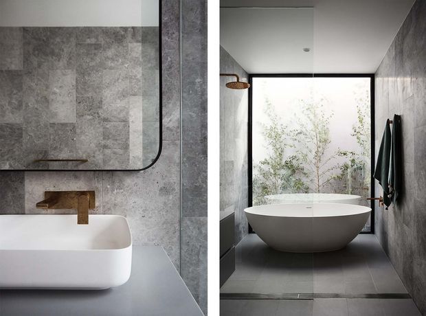 A refined contemporary bathroom featuring a freestanding white oval bathtub positioned before full-height glazing, grey stone-look wall and floor tiles, a wall-mounted timber-toned tap, a frameless black-edged mirror, and a dark-framed window overlooking greenery. A wall-mounted shower and towel hooks complete the minimalist fit-out.