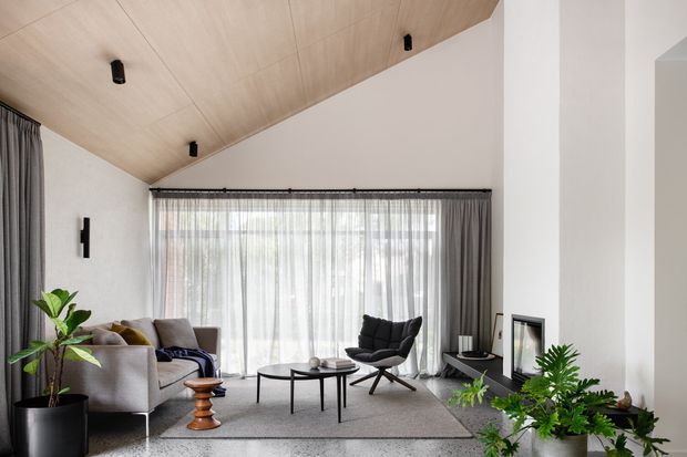 A bright, refined living room with a high sloped timber-lined ceiling, white walls, full-height sheer curtains, and a large glazed opening. The room features a neutral sofa and lounge chairs, a low round coffee table, indoor plants, a grey rug, recessed black lighting, and a minimalist fireplace set into the wall.