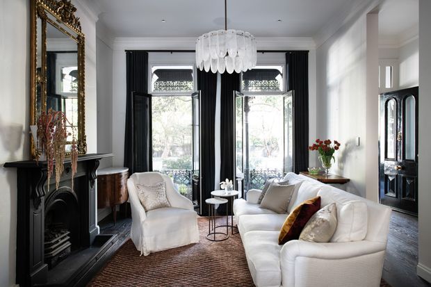 Elegant traditional living room with a black ornate fireplace and gilded mirror, tall French doors framed by black curtains, white upholstered seating, a glass pendant light, and dark timber flooring. The space combines refined period detailing with a bright, sophisticated neutral interior.
