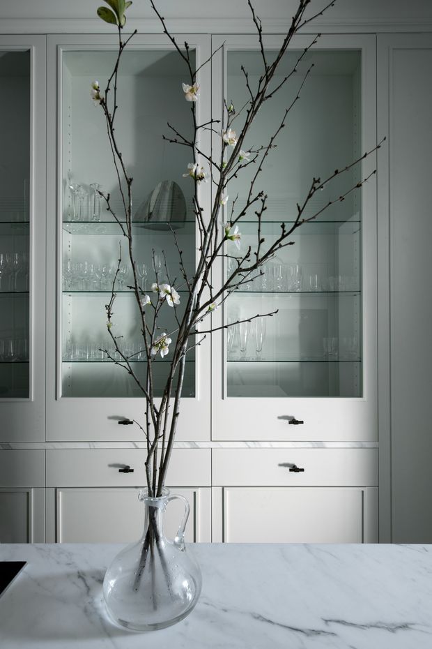 Close-up interior view of a refined white built-in display cabinet with glass-front doors, glass shelves, drawers, and dark hardware. The cabinet displays glassware, while flowering branches in a clear glass vase sit on a white marble island or countertop in the foreground.