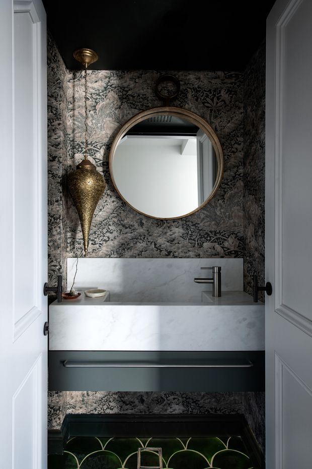 A compact, dramatically styled powder room viewed through open white paneled doors. The room features dark botanical patterned wallpaper, a black ceiling, a wall-mounted dark vanity with a pale marble benchtop and integrated backsplash, a sleek brushed-metal tap, a large round brass-framed mirror, and an ornate brass pendant light. Decorative dark green and black patterned floor tiles add further visual richness.