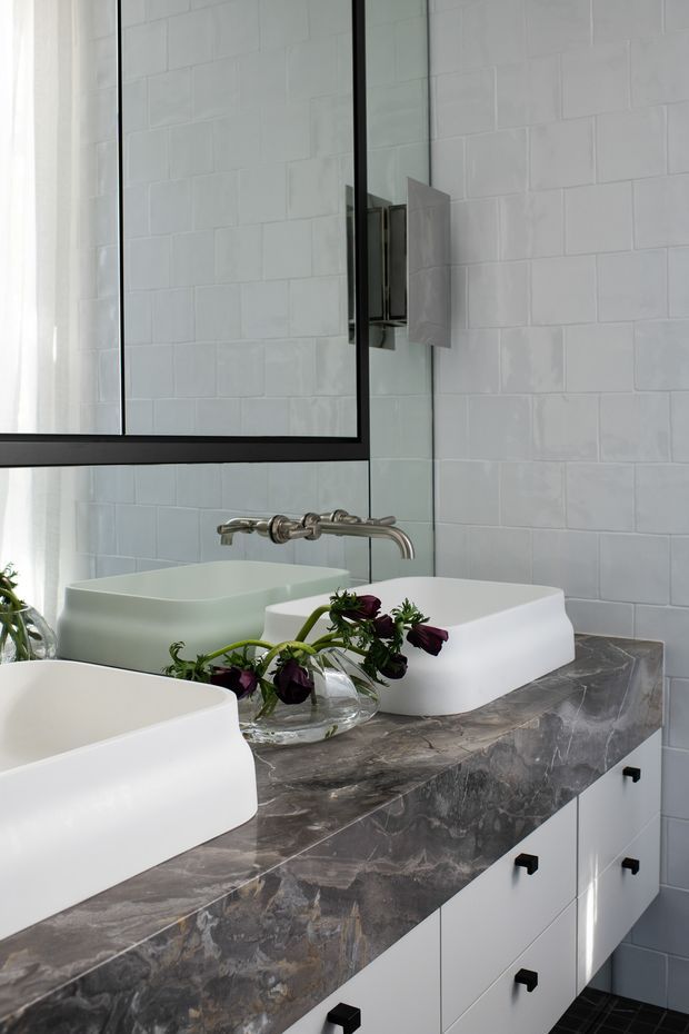 A refined contemporary bathroom vanity with two white vessel basins set on a dramatic dark grey veined stone benchtop. White rectangular wall tiles, a large black-framed mirror, brushed metal wall-mounted tapware, and white drawer cabinetry create a clean, monochromatic scheme with dark accents. A glass vase with greenery and deep purple flowers provides a small decorative contrast.