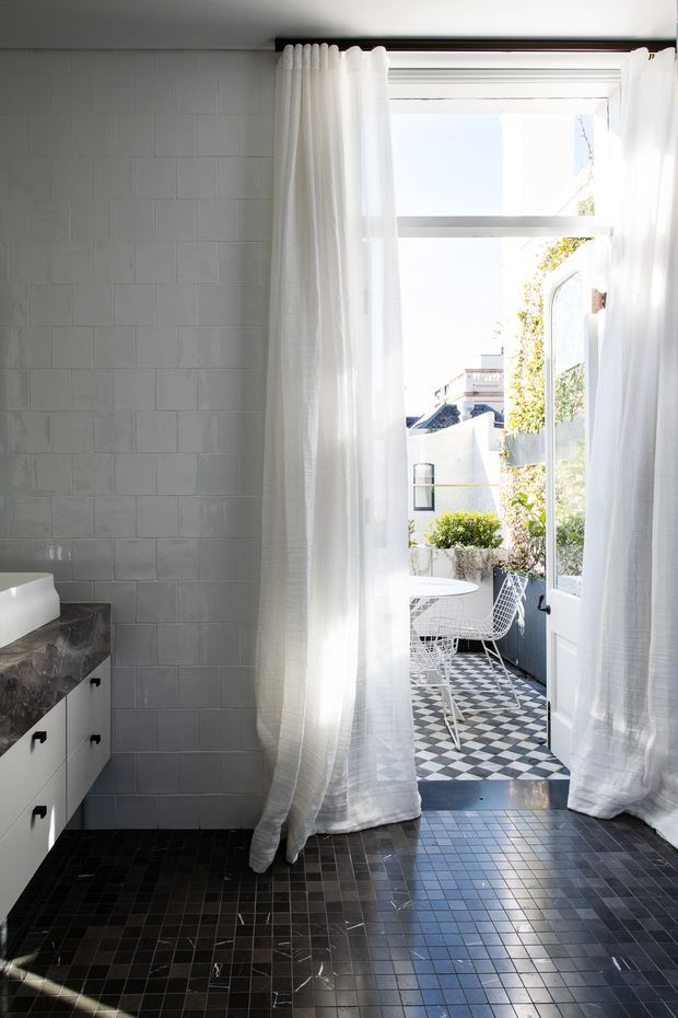 Bright contemporary bathroom with white rectangular wall tiles, dark small-format tiled flooring, a white vanity with dark stone benchtop, and a freestanding white bathtub partially visible at left. Sheer white curtains frame a glazed door opening to a small outdoor terrace with monochrome checkerboard paving and a wire chair. Strong natural light creates an airy indoor-outdoor connection.