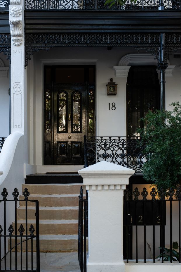 Front exterior view of a narrow, multi-storey terrace-style townhouse with an ornate black wrought-iron balcony, balustrades, gate and railings. The facade is painted white with decorative classical mouldings, carved column details, a black glazed entrance door, stone steps, and visible house numbers.