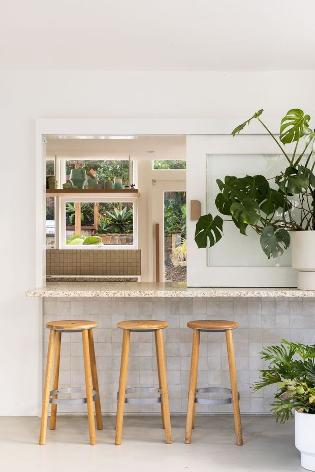 Bright, compact kitchen or entertaining area with a white-painted interior, a terrazzo-look countertop, pale square tiled backsplash, and three timber bar stools. A large internal opening and glazed door connect visually to a leafy garden, while substantial potted tropical plants add a natural accent. The space has a clean, relaxed contemporary feel.