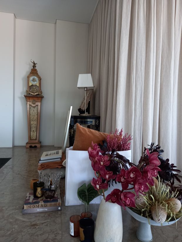 Elegant, softly lit living room with pale panelled walls, full-height sheer curtains, a marble-look floor, an ornate antique grandfather clock, a classic table lamp, and a console or side table. A coffee table in the foreground is styled with books, glassware, candles, and a large floral arrangement in sculptural vases. The room has a refined, traditional character with contemporary styling.