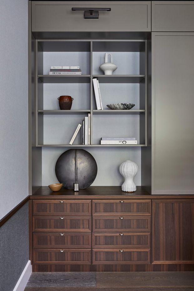 Close-up interior view of a sophisticated built-in storage and display unit. Dark timber-grain cabinetry with multiple drawers and a cupboard is paired with a grey open shelving niche, displaying books and sculptural decorative objects. The composition has a refined, moody, mid-century-inspired character with dark wood flooring and a textured grey wall.