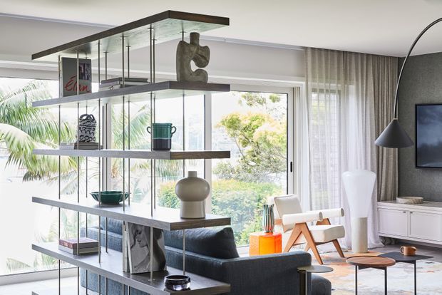 Bright contemporary living room with a dramatic floor-to-ceiling glazed opening, sheer curtains, a dark textured feature wall, and a freestanding open shelving divider displaying decorative objects. The room includes a dark sofa, sculptural white lounge chair, timber-framed armchair, nesting coffee tables, floor lamp, and low white media cabinetry. Lush greenery is visible outside through the glazing.