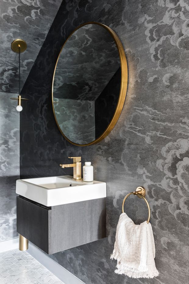 A compact, dramatically styled powder room featuring a wall-mounted white basin with dark timber-look vanity cabinetry, brushed gold tapware and hardware, an oval gold-framed mirror, and a slim pendant light. The walls are covered in dark grey patterned wallpaper, complemented by small pale geometric floor tiles and a textured white hand towel.