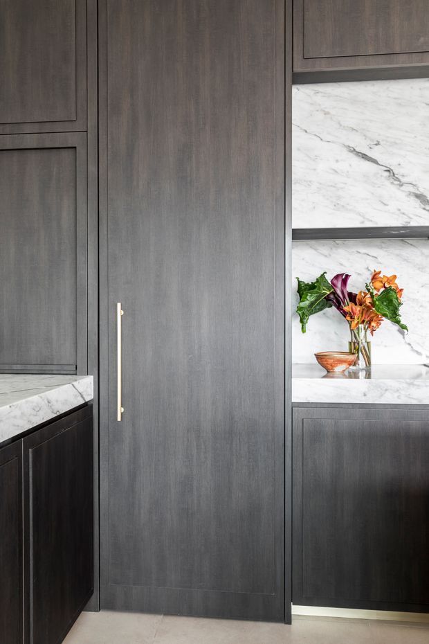 Close-up interior view of a refined dark timber kitchen or butler's pantry, featuring full-height paneled cabinetry, a tall integrated door with a slim brass handle, and white veined marble benchtop and wall surfaces. A small floral arrangement adds colour on the open marble shelving.
