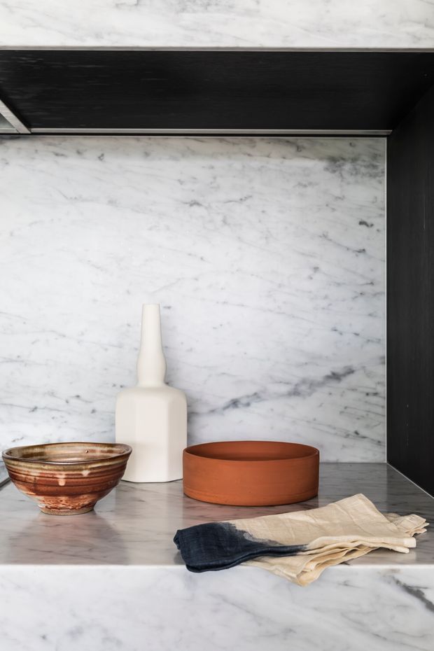 Close-up of a refined marble-lined built-in display or kitchen shelving niche. White veined marble forms the back, shelf, and lower counter, framed by dark black cabinetry or panels. Minimal ceramic vessels and bowls in white, terracotta, and brown sit alongside folded neutral and navy textiles, creating a restrained, high-contrast vignette.