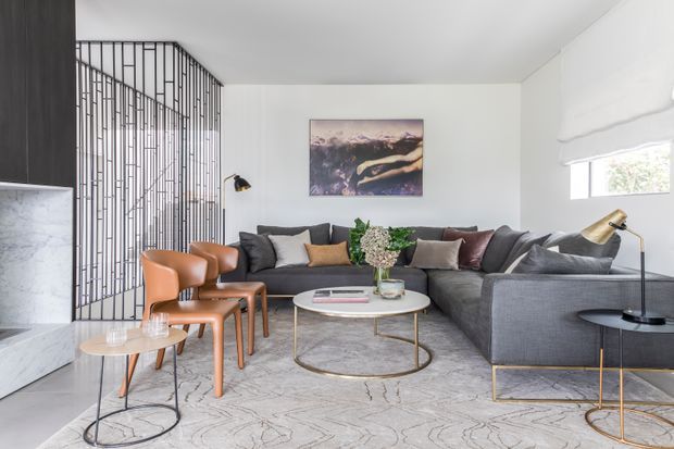 Bright, contemporary living room with a large grey sectional sofa, sculptural leather dining-style chairs, round marble coffee table, and slim brass-toned furniture frames. A black metal-framed open stair or screen structure forms a strong architectural feature at left, while a soft neutral rug, white walls, contemporary artwork, and layered table lamps complete the refined interior.