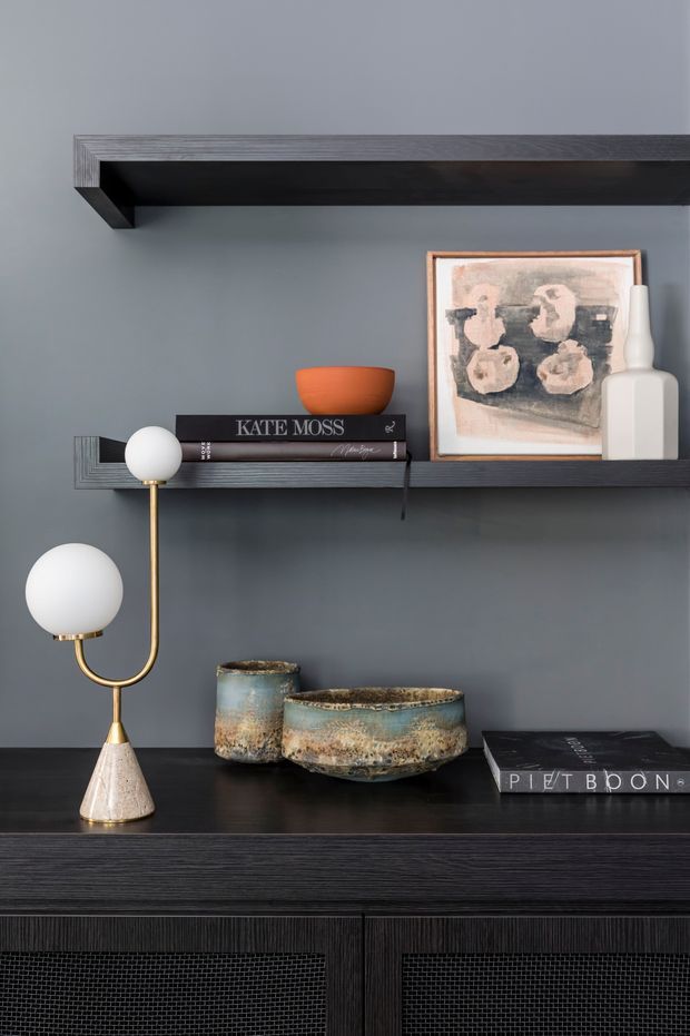 Close-up interior vignette featuring dark textured built-in shelving and cabinetry against a muted grey wall. The shelves display books, framed artwork, sculptural white vessels, ceramic bowls, and a decorative brass-and-white lamp. The composition is refined and dark-toned, with a contemporary decorative styling.