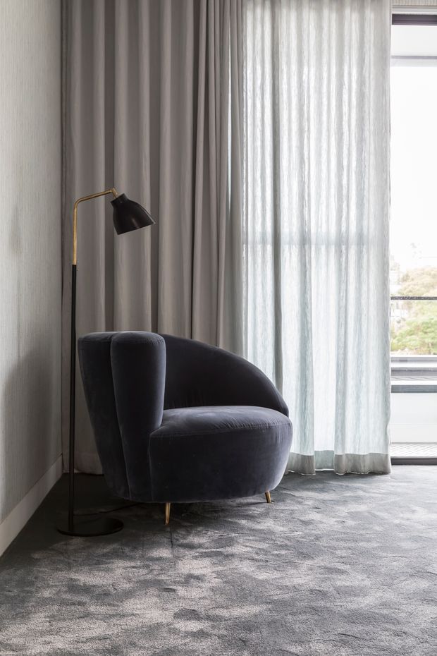 A softly lit contemporary seating corner with a dark charcoal upholstered lounge chair, slim black-and-brass floor lamp, full-height grey drapery and sheer curtains, textured grey carpet, and a large glazed opening overlooking greenery. The scene has a refined, quiet hotel or residential interior feel.
