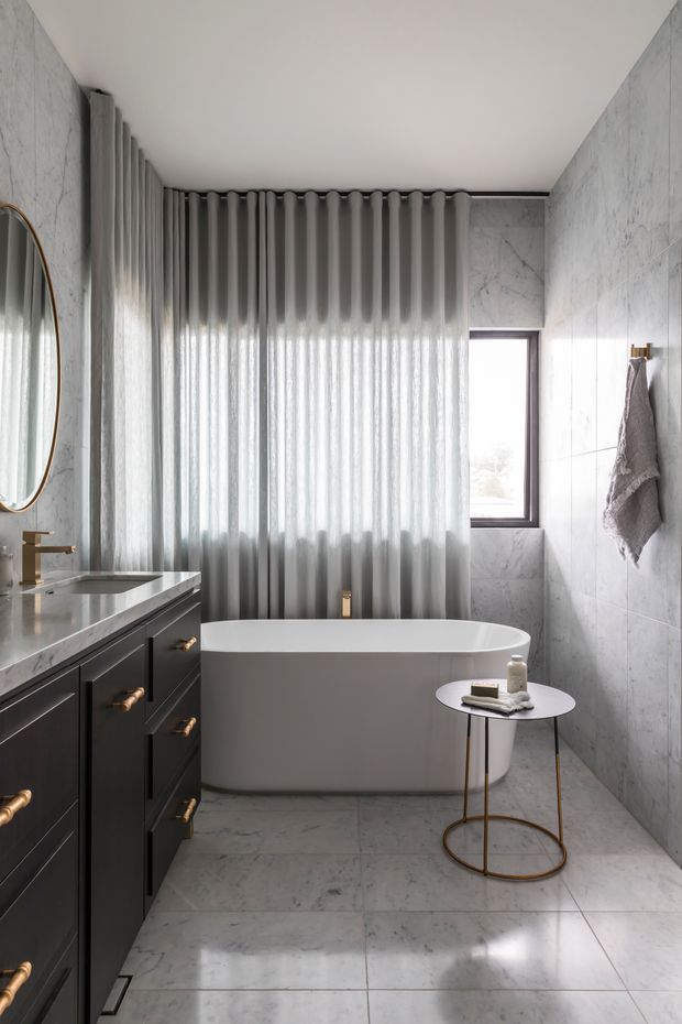 A refined contemporary bathroom featuring a freestanding white bathtub beneath full-height sheer grey curtains, large white-grey marble-look wall and floor tiles, a dark charcoal vanity with brass hardware, a stone benchtop and integrated basin, a brass tap, an oval wall mirror, and a small round side table. Natural light enters through a narrow window, creating a calm, luxurious monochromatic atmosphere.