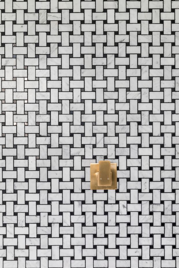 Close-up of a decorative bathroom wall featuring a repeating basket-weave mosaic pattern of white and light grey marble-look rectangular tiles with dark grout lines. A small brass/gold wall control or fixture is mounted near the centre.