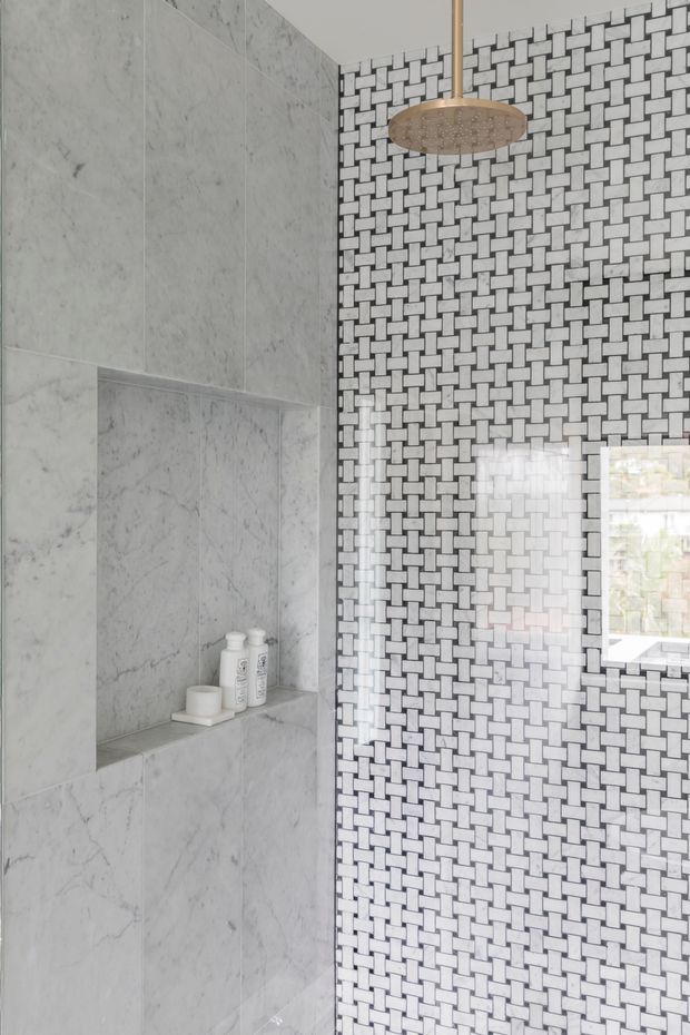 A refined bathroom shower area featuring pale marble-look wall slabs with an integrated recessed niche, contrasted by a full-height black-and-white woven basketweave mosaic tile feature wall. A round brushed-gold rainfall showerhead and a small window complete the space.