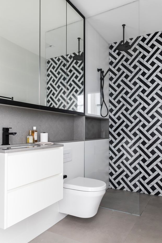 Contemporary bathroom with a floating white vanity, integrated basin, wall-mounted toilet, large mirrored cabinet, matte black tapware and shower fittings, and a dramatic black-and-white herringbone tiled shower feature wall. Large grey floor tiles and grey wall surfaces create a clean monochromatic scheme.