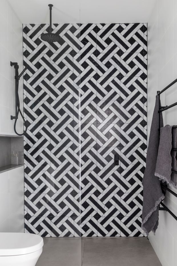 Compact contemporary bathroom featuring a walk-in shower enclosed by clear glass, a bold black-and-white herringbone tile feature wall, black shower fittings, a wall-mounted towel rail with dark towels, white wall tiles, and large grey floor tiles. A partial toilet and vanity are visible at the lower and left edges.