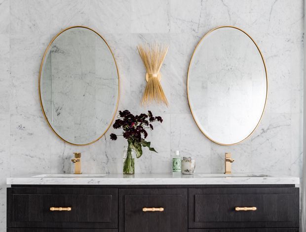 A refined double-vanity bathroom featuring a dark timber-look floating vanity with brass handles, a white marble-look wall and benchtop, two gold-framed oval mirrors, and matching brass tapware. Minimal decorative styling includes a central dried botanical wall feature and a small vase.