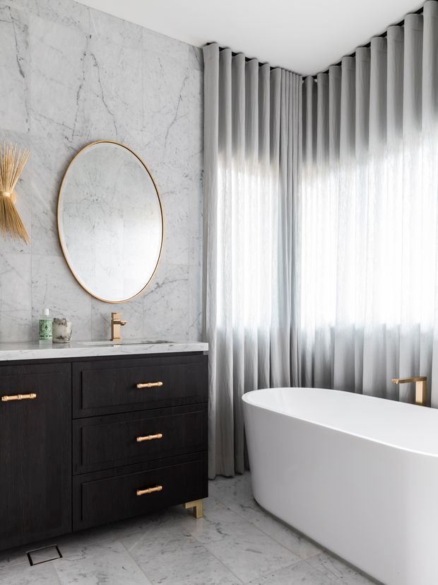 A refined modern bathroom featuring a freestanding white bathtub beside full-height sheer grey curtains. The space has large veined marble-look wall and floor surfaces, a dark timber vanity with brass handles and legs, a brass tap, and a large round mirror with a brass frame. Soft daylight creates a bright, luxurious atmosphere.