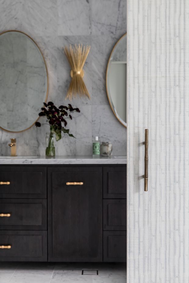 A refined bathroom vanity vignette featuring dark timber cabinetry with brass pulls, a pale marble benchtop and full-height marble wall finish. Two large oval mirrors, a sculptural brass wall light, and a partially visible light textured door or screen create a luxurious, layered composition.