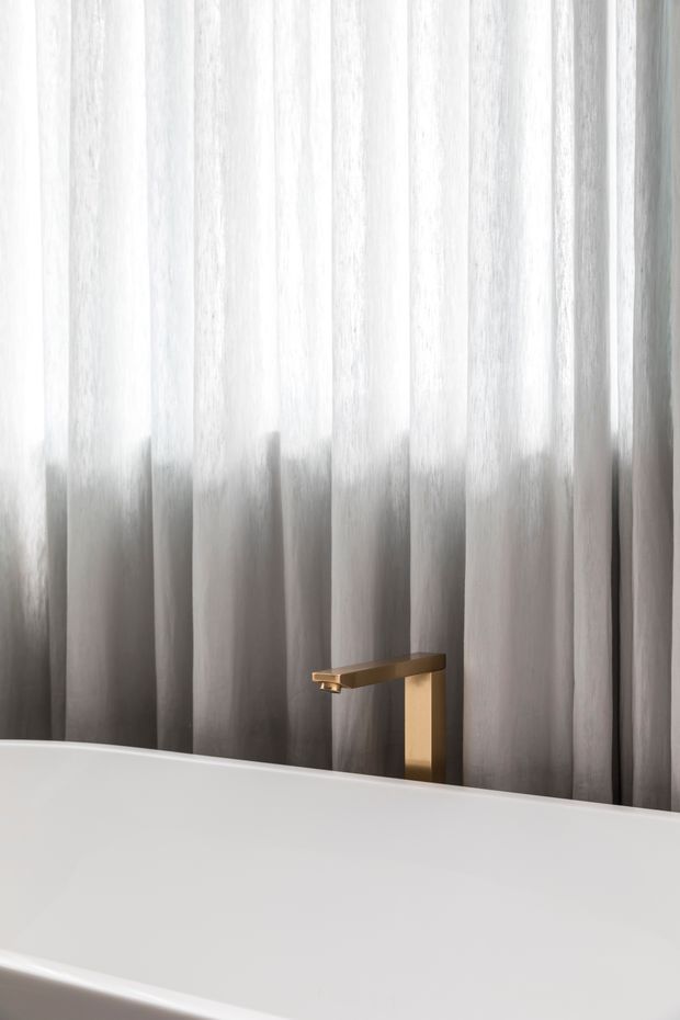 Close-up of a minimalist bathroom featuring a white freestanding bathtub, a wall-mounted or deck-adjacent brushed gold/brass tap, and floor-to-ceiling sheer grey curtains with soft vertical folds behind the tub. The composition is quiet, refined, and softly lit.