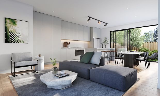Bright open-plan contemporary living and dining space with a compact white kitchen, integrated cabinetry, dark grey sectional sofa, sculptural coffee table, and a dining area opening to a landscaped outdoor space through large black-framed glazed doors. The room has a clean neutral palette, track lighting, timber flooring, and soft natural light.