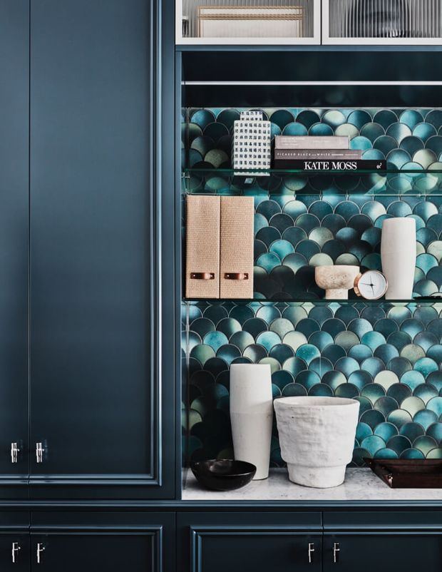 Close-up of a dark navy built-in storage cabinet with framed shaker-style doors and drawers. The open display section has a striking glossy scalloped mosaic tile backdrop in teal, turquoise, blue, and charcoal, with glass shelves holding books, storage boxes, ceramics, a clock, and decorative objects. The composition is highly detailed and moody, with contrasting pale stone or marble shelving surfaces and metallic hardware.