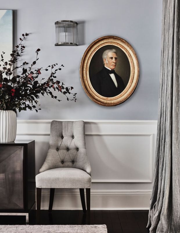 A refined interior vignette featuring a tufted upholstered accent chair beside a dark timber cabinet, with white wainscoted wall panelling, a framed oval portrait, a wall sconce, draped curtains, and a decorative branch arrangement. The palette is soft grey, white, black, and warm wood tones, creating a formal, classic atmosphere.