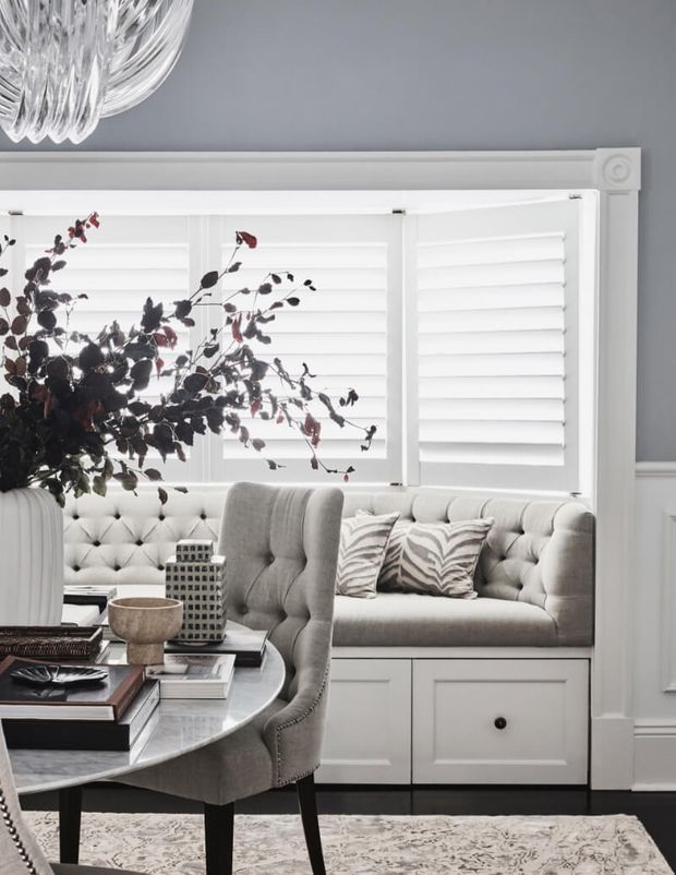 Elegant traditional sitting and dining area with a built-in tufted window seat, white plantation shutters, grey walls, decorative wall moulding, and a round marble-topped table surrounded by upholstered seating. A crystal chandelier and patterned rug add refined detail, while muted grey, white, and black tones create a polished classic interior.