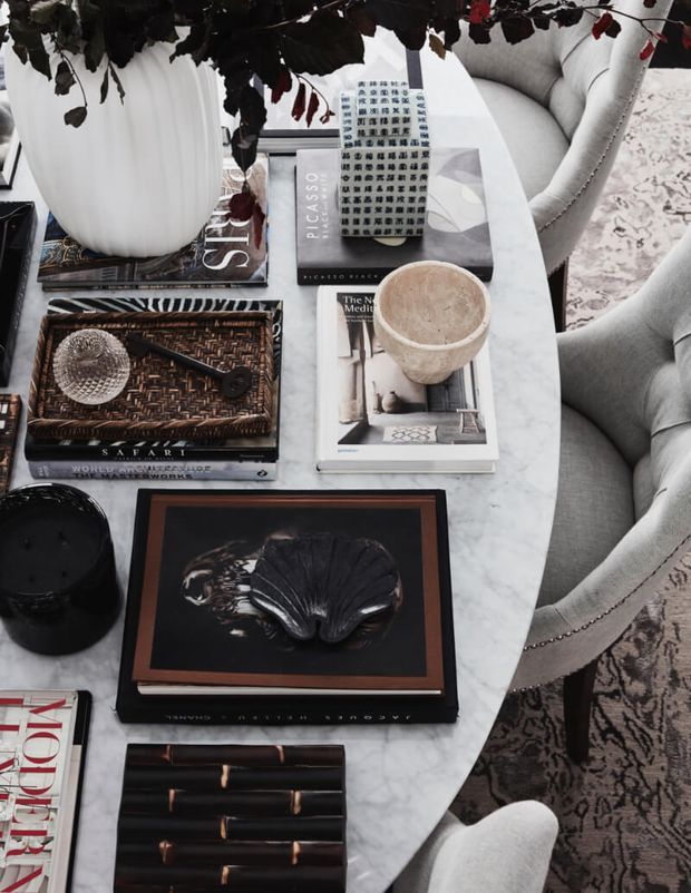 Close-up view of a refined living-room seating area centered on an oval white marble coffee table styled with art and design books, a sculptural black object, ceramic cup, decorative tray, and large white vase with dark foliage. Curved light upholstered lounge chairs and a patterned rug surround the table. The palette is monochromatic and sophisticated, with black, white, grey, and dark brown accents.