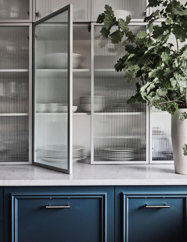Close-up interior detail of a refined kitchen or dining storage area, featuring dark navy-blue base cabinetry with framed drawer fronts, a pale stone benchtop, and tall white-framed reeded glass cabinet doors. White dishes are displayed on open shelving behind the glass, alongside a large leafy potted plant. The composition has a sophisticated, high-contrast contemporary character.