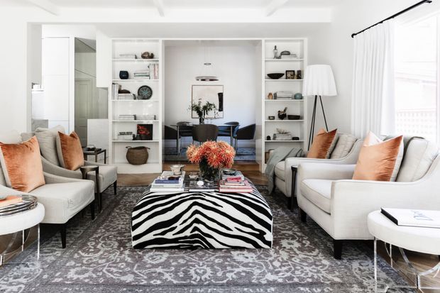 Bright, refined living room with white built-in shelving framing a dining area, paired upholstered armchairs and sofas, a patterned area rug, and a bold zebra-patterned coffee table ottoman. Tall white curtains and a coffered ceiling create an airy, polished atmosphere, with warm rust and orange cushions adding contrast.