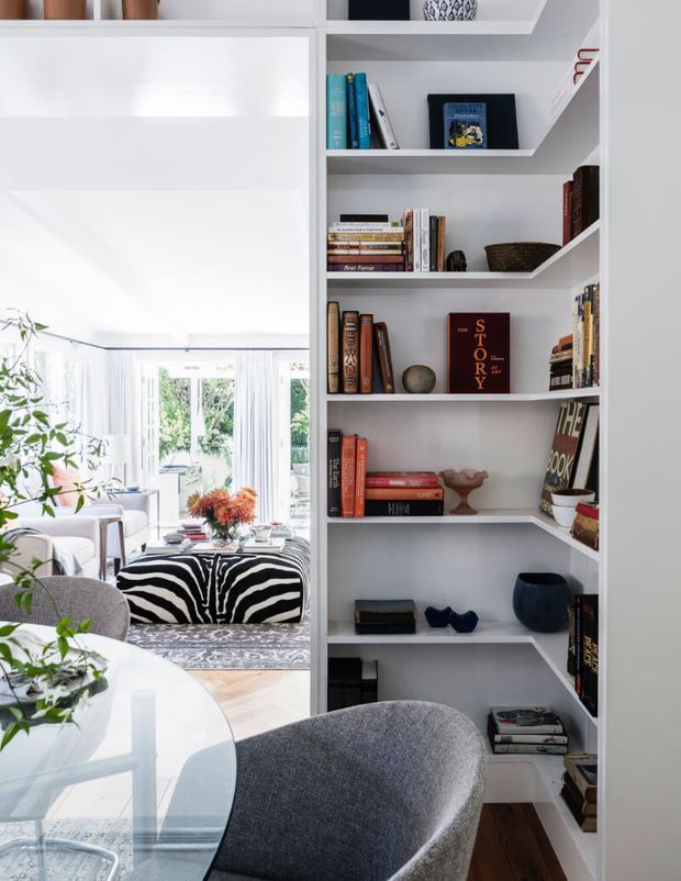 Bright contemporary interior featuring a built-in white shelving unit filled with books and decorative objects, opening onto a light-filled living room. A round glass dining table and upholstered grey chair occupy the foreground, while the adjoining lounge includes a zebra-patterned ottoman, pale seating, abundant glazing, indoor plants and timber flooring.