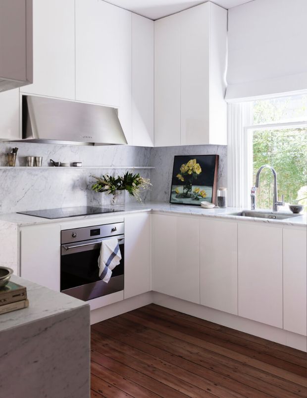 Bright, compact contemporary kitchen with white flat-panel cabinetry, a marble-look splashback and benchtops, integrated stainless-steel oven and rangehood, and a deep stainless-steel sink beneath a window. Dark timber flooring contrasts with the predominantly white fit-out, while a small peninsula and minimal decorative styling complete the space.