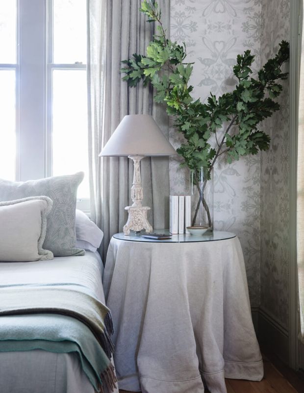 A softly lit, refined sitting-room corner featuring a round side table draped in a pale fabric cloth, a decorative white lamp, books, and a tall leafy branch arrangement in a glass vase. A pale upholstered sofa with layered cushions and throws sits beside a large window, while patterned grey wallpaper and full-length curtains create an elegant, calm backdrop. Timber flooring is visible at the base.