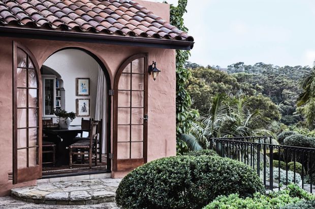 A Spanish-style stucco building exterior with a terracotta barrel-tile roof and arched timber-framed French doors opening onto a small stone terrace. A furnished interior is visible through the doorway, while manicured shrubs, a black metal balustrade, and lush tropical vegetation frame a wooded outlook.
