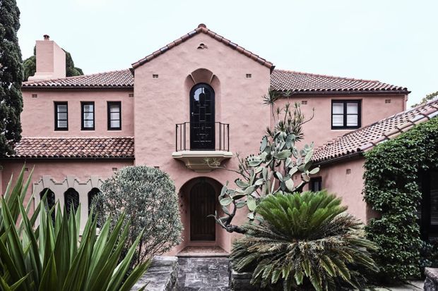 Exterior view of a pink stucco, Mediterranean-inspired residence with a symmetrical central gabled entry, arched dark timber door, small balcony, terracotta tiled pitched roofs, chimney, and dark-framed windows. Mature drought-tolerant landscaping, including palms, agave, yucca, and cycads, frames the facade.