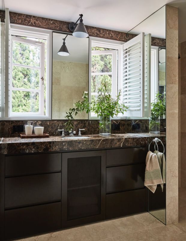 Elegant bathroom vanity featuring dark brown stone surfaces, dark built-in cabinetry, large mirrored panels, white-framed windows, and white plantation shutters. The space has a refined, moody yet naturally lit atmosphere, with a marble-clad wall and floor and classic wall-mounted pendant lights.