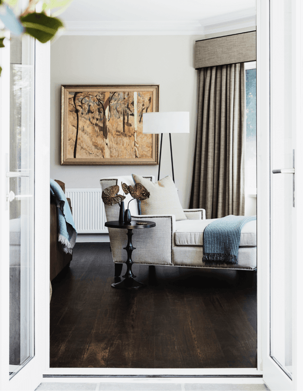 A refined living room viewed through open white-framed glazed doors. The room features dark timber flooring, pale grey walls with white ceiling mouldings, full-height taupe curtains, a tailored cream upholstered sofa and armchair, a black-and-white floor lamp, a small black pedestal table, framed abstract artwork, and blue textured throws. The styling is elegant, layered, and classic with strong contrast between the dark floor and light furnishings.