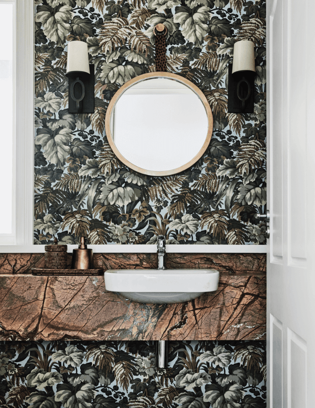A compact, highly styled bathroom vanity viewed straight on. A round timber-framed mirror is centred against dramatic botanical wallpaper, flanked by black wall sconces. Below is a white wall-mounted basin with a chrome tap, set into a richly veined brown stone vanity surround. White trim and a partially open white door frame the scene.