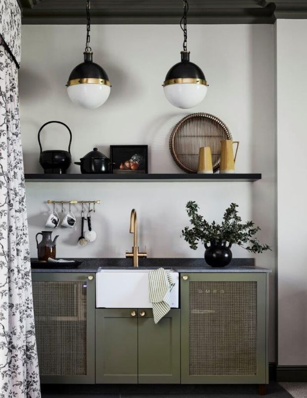 A compact, highly styled kitchen with muted green shaker cabinetry, woven mesh-front cupboard doors, a white farmhouse sink, dark benchtops, and brass tapware. Open black shelving displays black ceramics, framed artwork, woven decor, and yellow vessels. Two black-and-brass pendant lights hang beneath a dark exposed-beam ceiling, while a patterned curtain frames the left side.