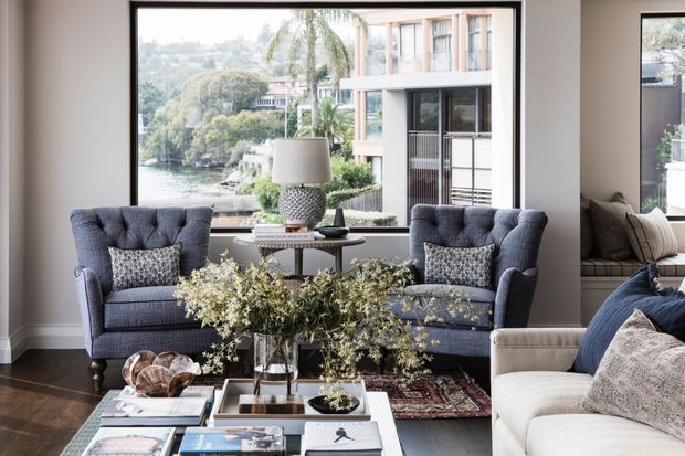 Refined living room with two tufted navy-blue armchairs facing a low coffee table, a pale upholstered sofa in the foreground, layered books and decorative accessories, and a large picture window overlooking lush waterfront surroundings and neighbouring architecture. The room features dark timber flooring, neutral walls, substantial window trim, and a composed traditional-contemporary styling.
