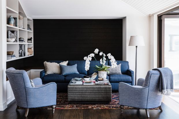 A refined living room with a deep navy-blue sofa, two upholstered blue armchairs, a textured dark coffee table, and a patterned area rug. Built-in white shelving displays books and decor along one side, while a dark horizontal timber feature wall anchors the seating area. White slatted ceilings, light walls, timber flooring, and large glazing create a bright, balanced interior.